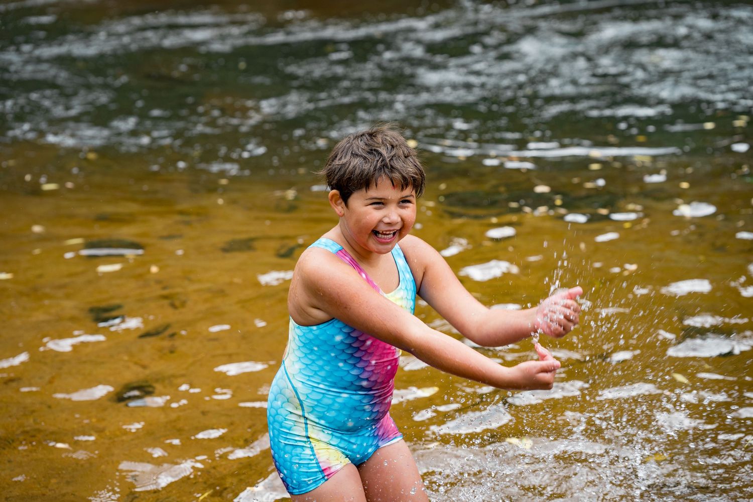 Child in blue swimsuit plays and splashes in shallow water during summer fun at the creek.