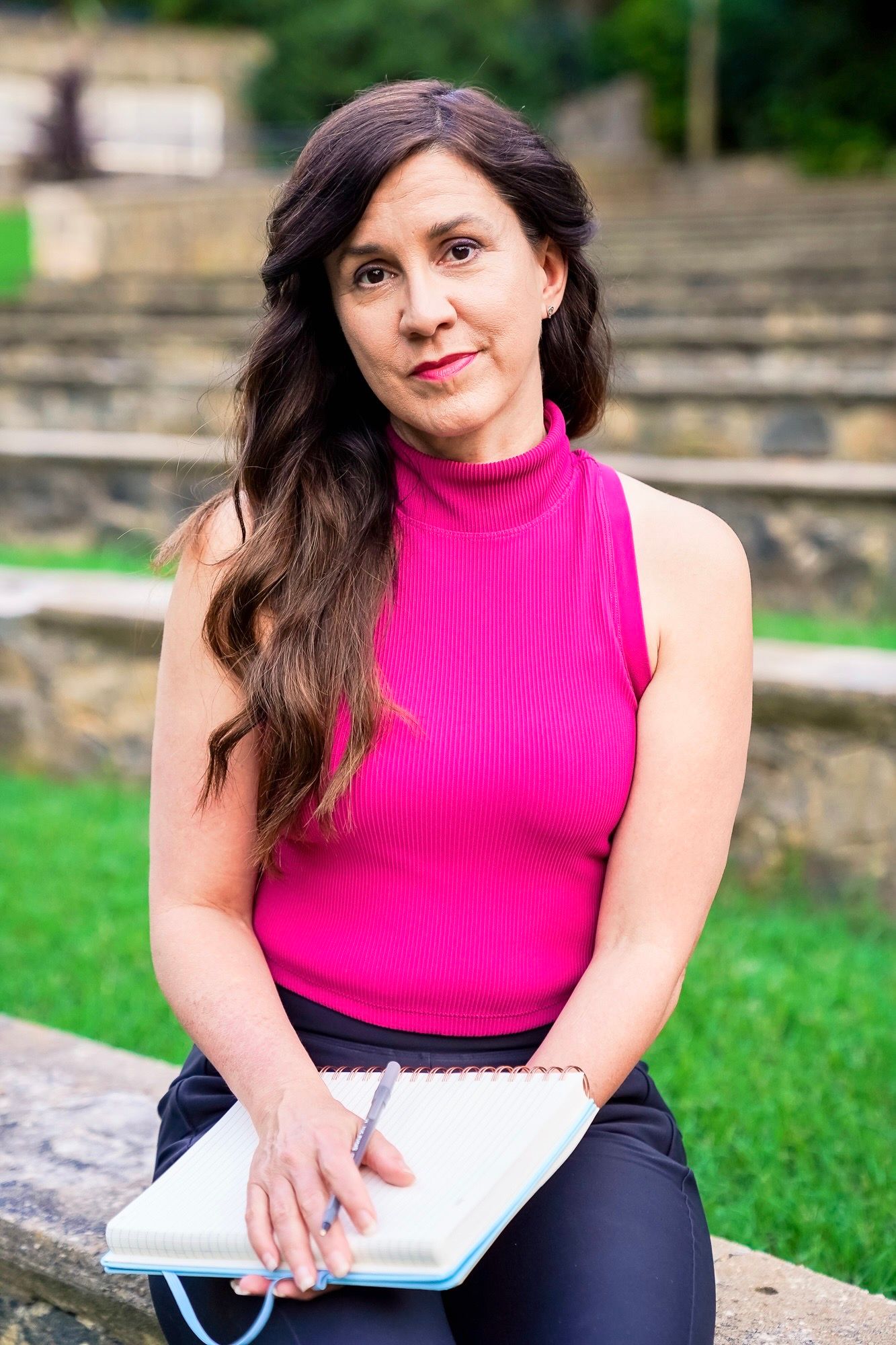 Person in bright pink sleeveless top and dark pants sits outdoors writing in notebook on stone steps.