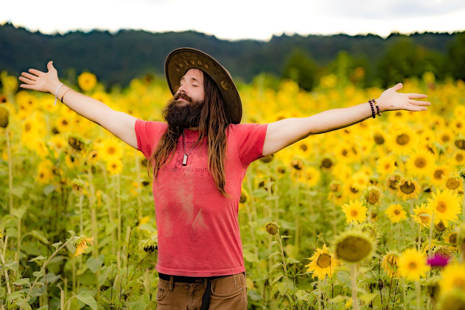 Bearded man with dreadlocks in red shirt and hat stands with arms outstretched in a vibrant sunflower field.