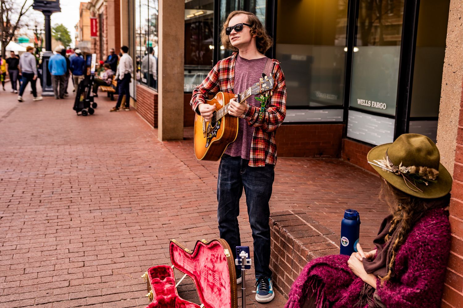Young street musician playing acoustic guitar on a brick sidewalk while a seated woman in a hat watches nearby.