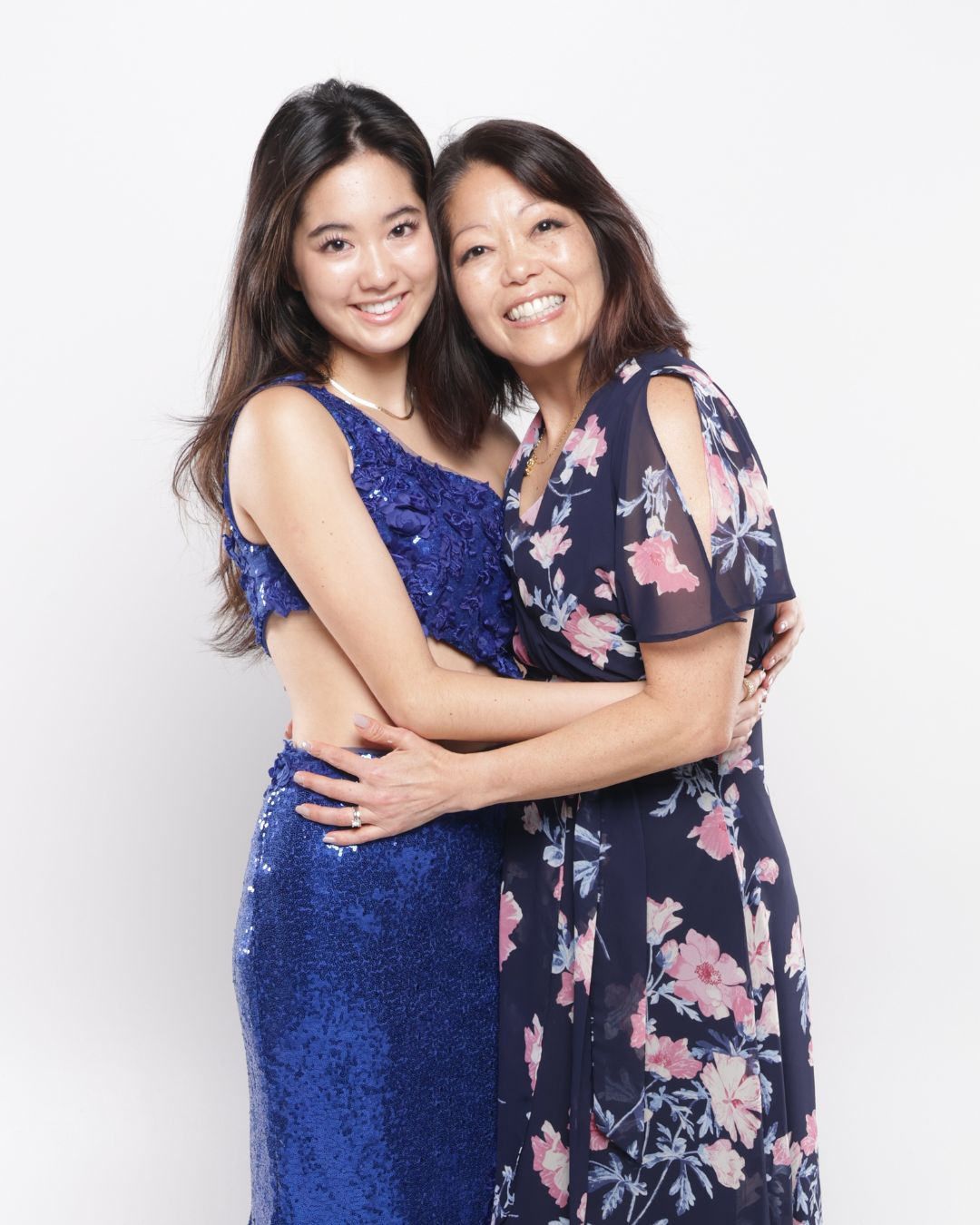 Two smiling women in blue floral dresses embrace warmly against a white background in a portrait photo.