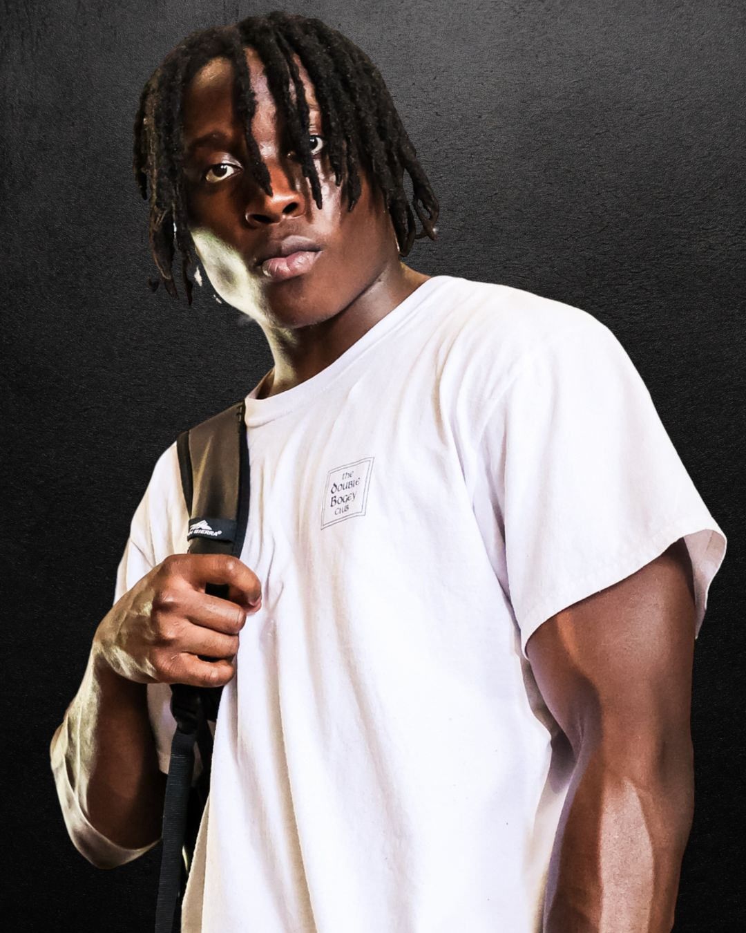 Young man with braids wearing a white t-shirt and carrying a backpack against a dark background.