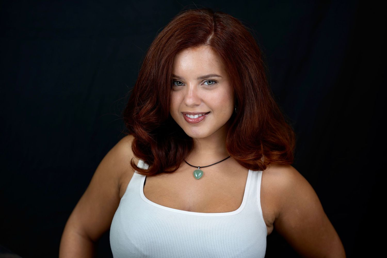 Young woman with red hair wearing a white tank top and teal necklace, smiling against a dark background.