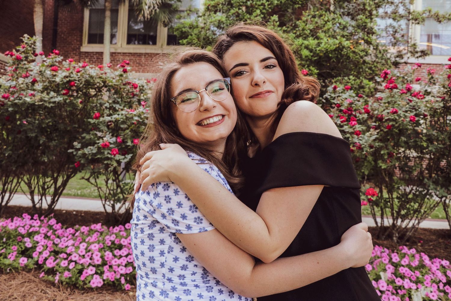 Two young women smiling and hugging outdoors near blooming pink flowers and green shrubs.