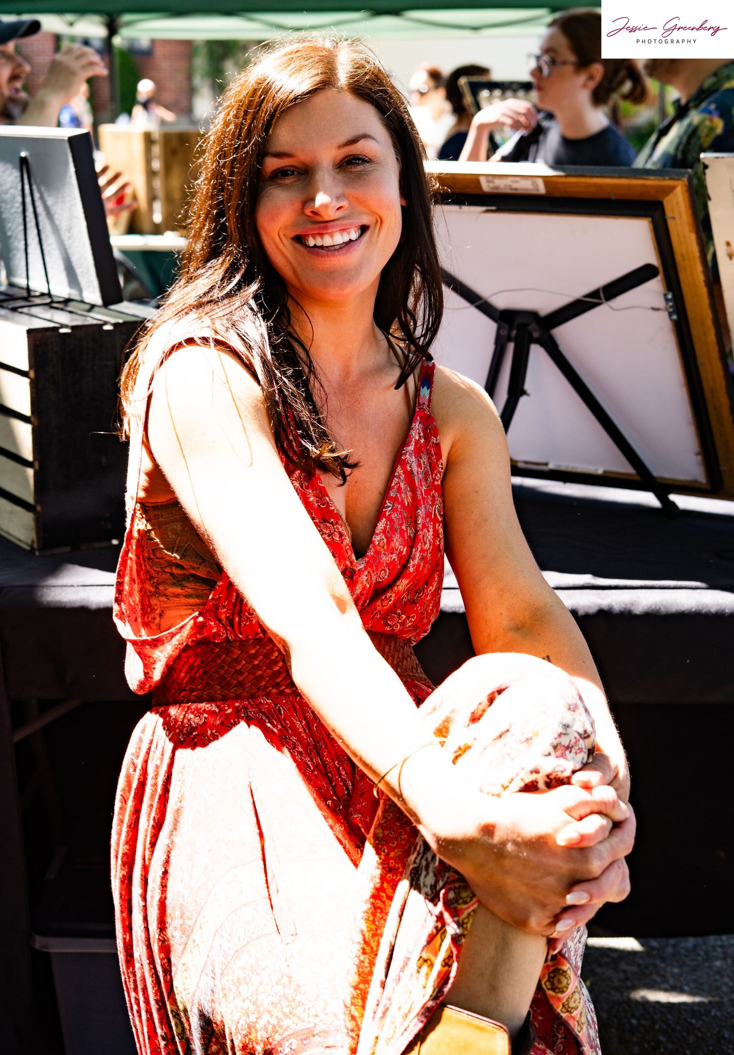 A smiling woman in a red floral dress sits outdoors at a sunny art market, with framed artwork visible behind her.