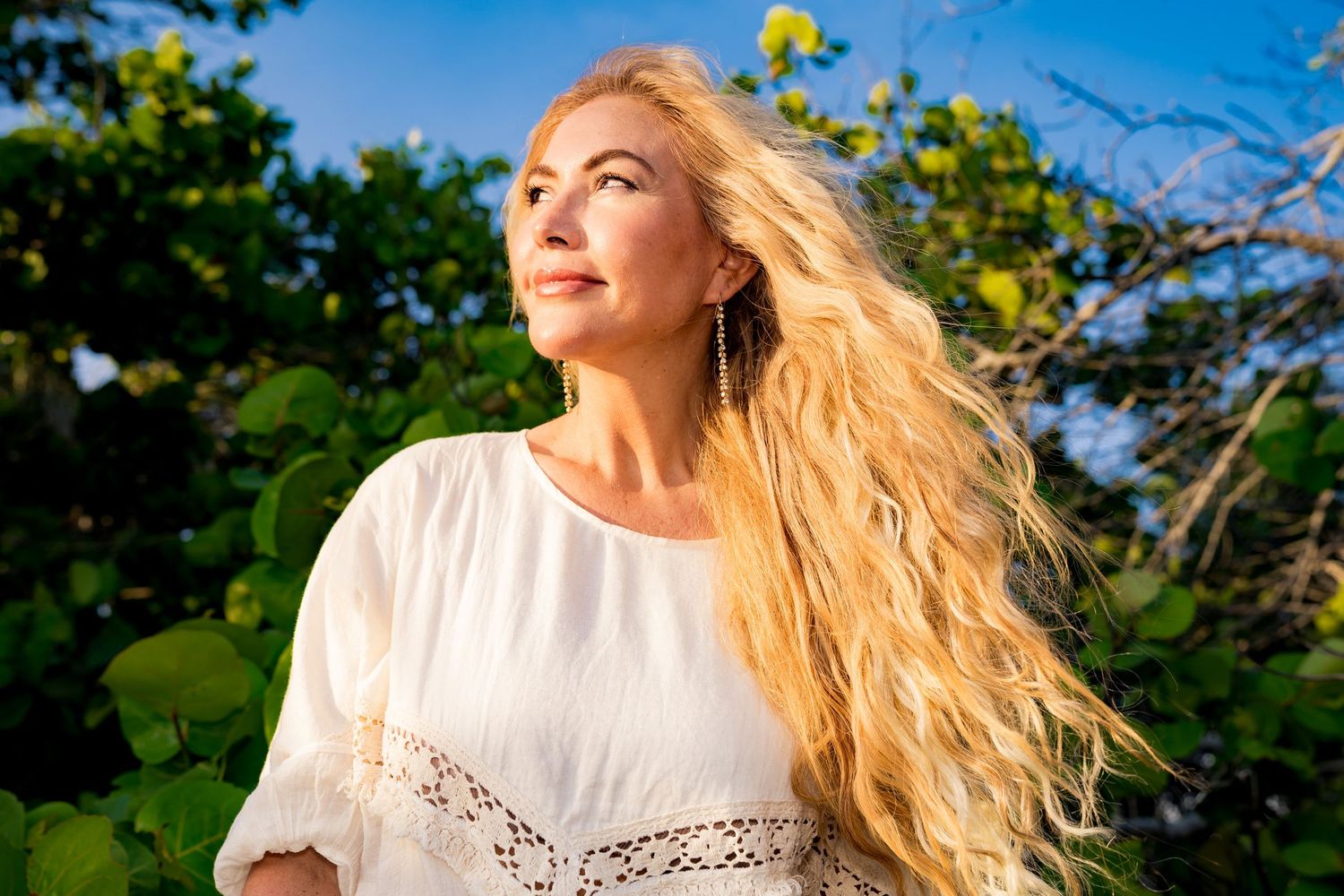 A photo of a blonde woman with flowy hair looking out into the distance, she is in the warm light with green behind her.