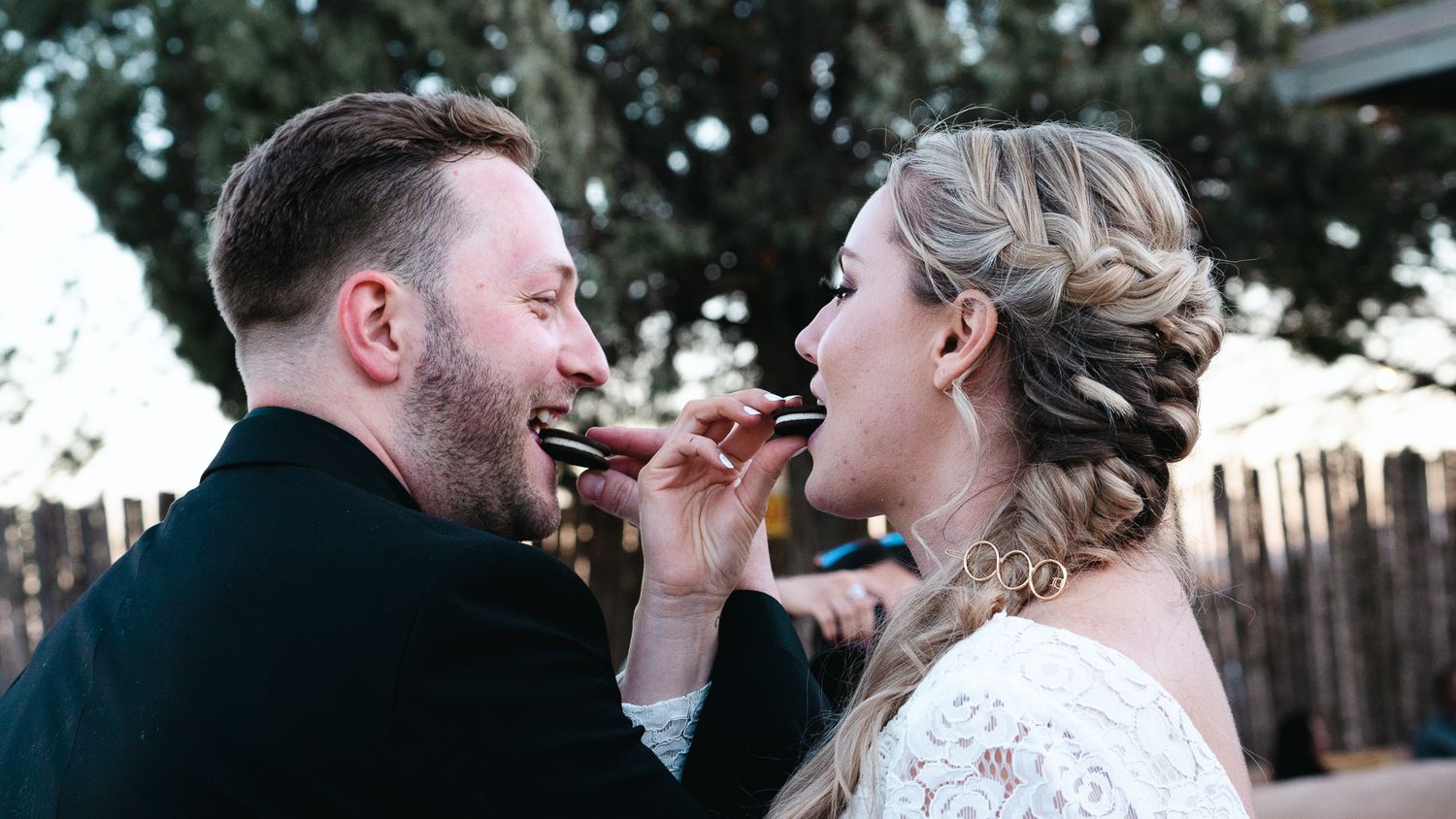Bride and groom sharing a bite of food outdoors at their wedding reception, smiling joyfully together.
