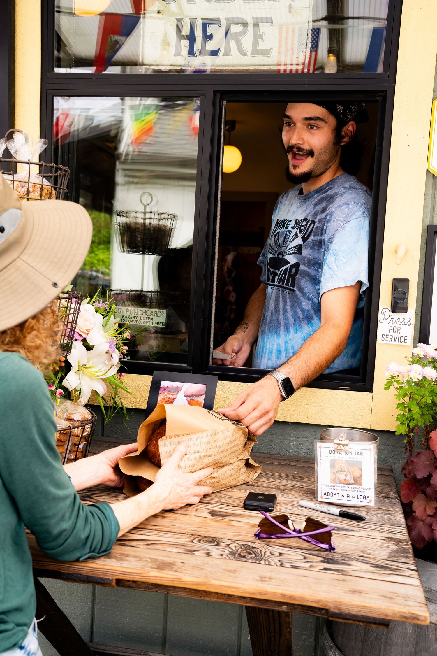 Young man with mustache in tie-dye shirt hands brown bag to customer at outdoor service window.