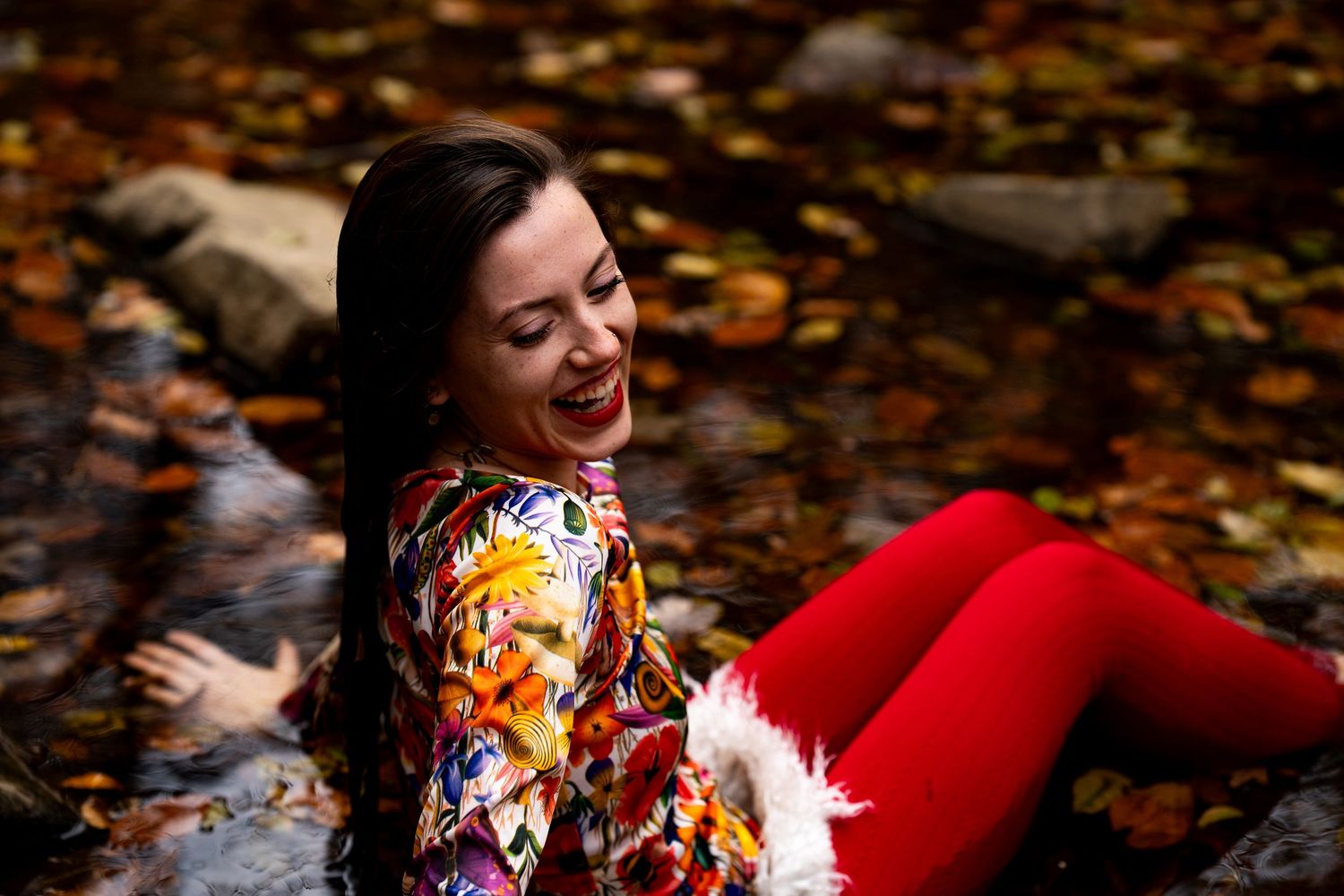 A person wearing a colorful floral dress lies on autumn leaves while laughing joyfully in a park setting.
