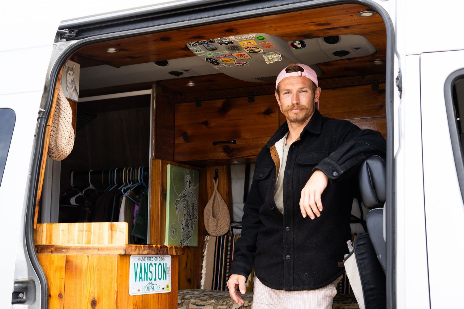 Man standing in open van conversion with wooden interior, hanging clothes, and a custom VANSTON license plate.