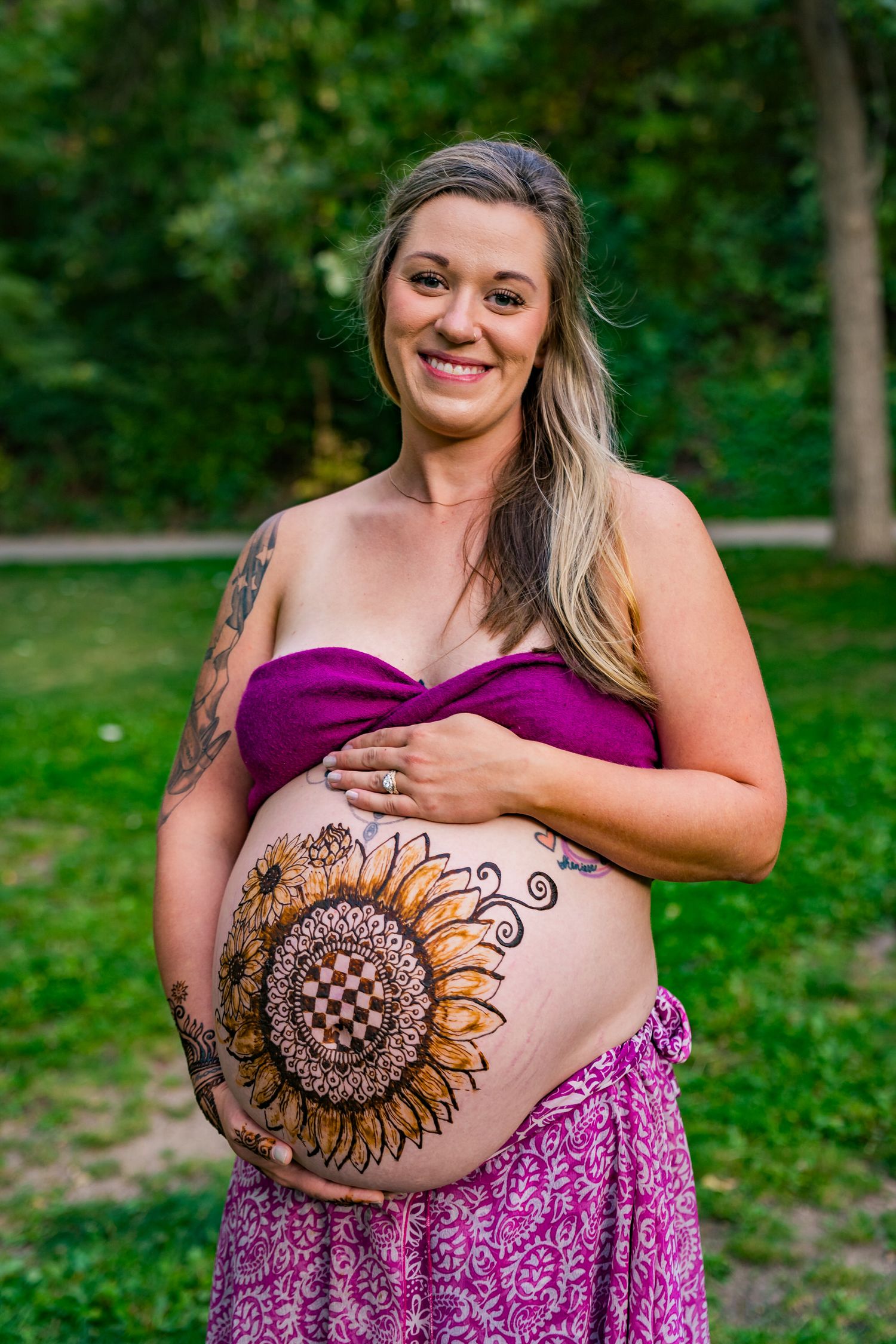 Smiling pregnant woman with henna sunflower belly art wearing purple bandeau top outdoors in green park setting.
