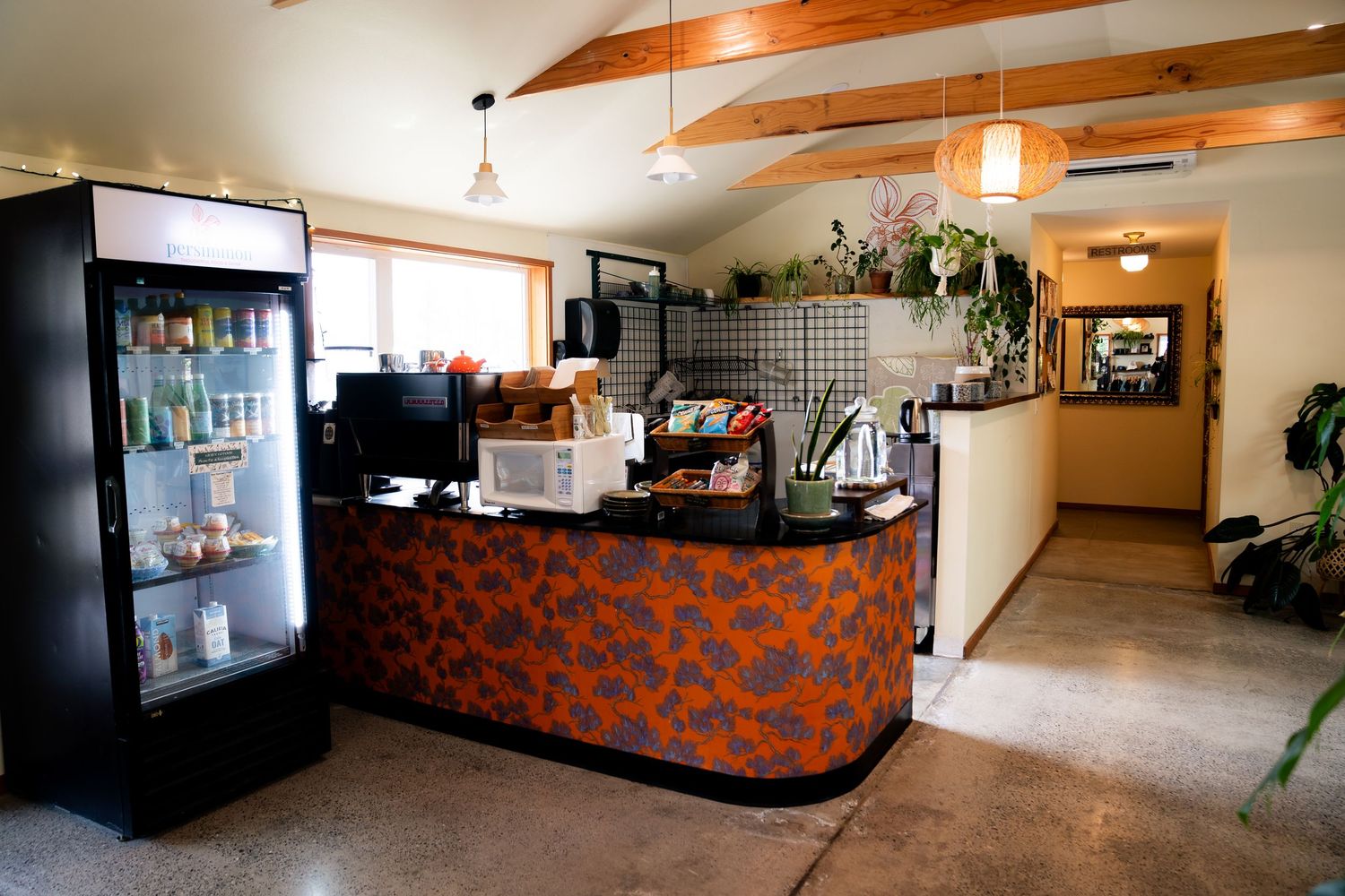 Cozy cafe interior with orange floral counter, glass refrigerator, and plants under vaulted wood beam ceiling.