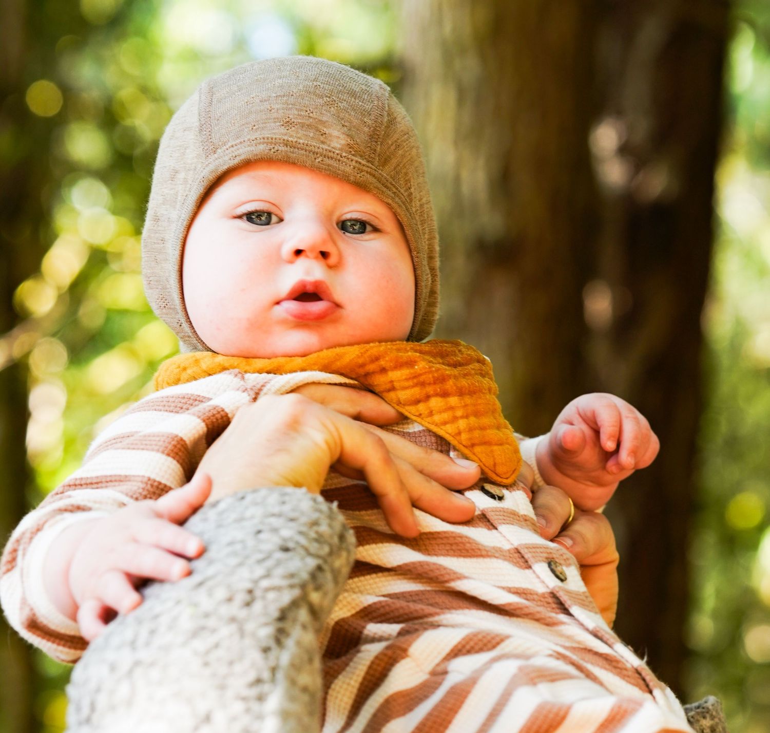 A cute baby wearing a beige hat and striped outfit with an orange bib is held up outdoors among trees.