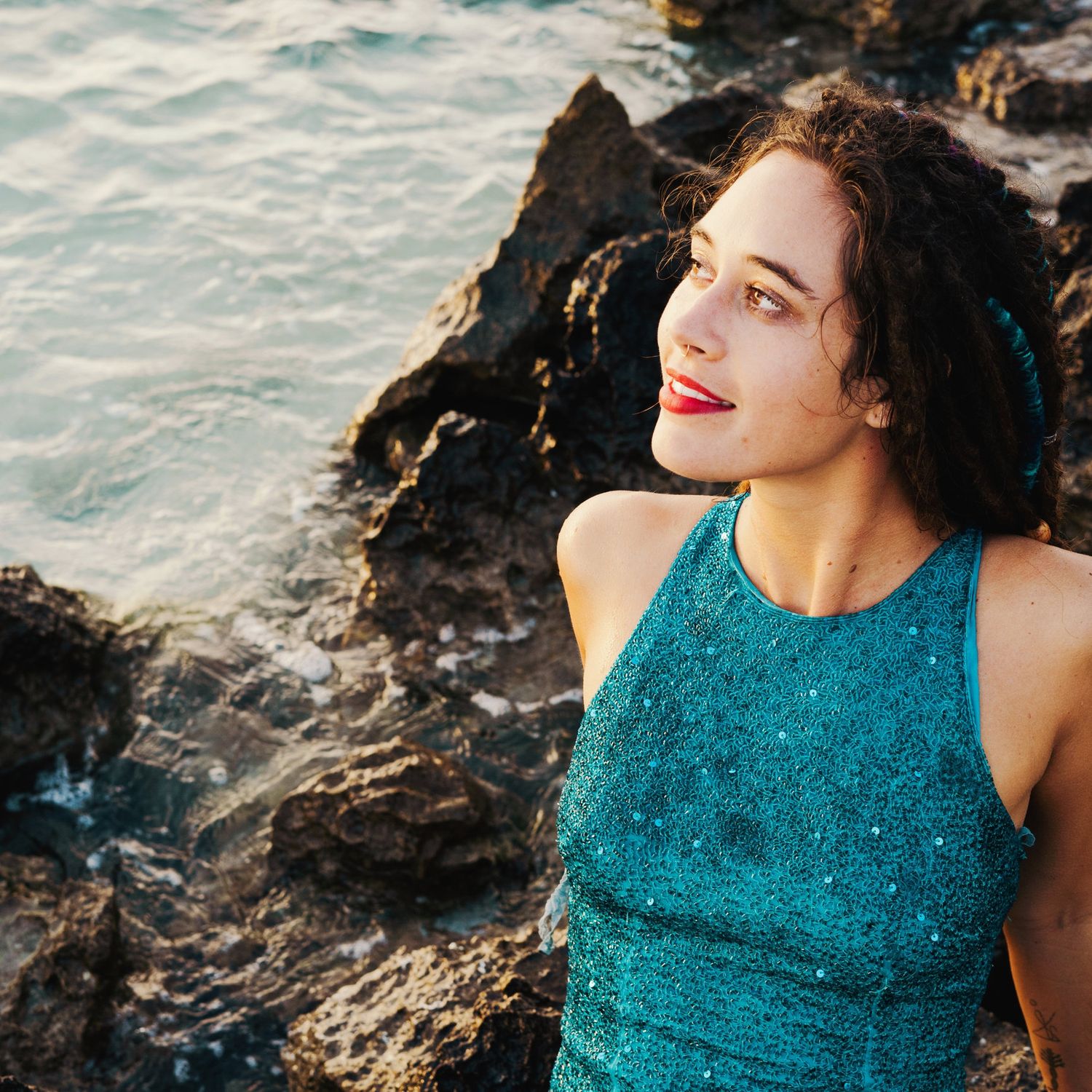 A photo of a woman wearing a light blue sequined dress and looking to the side sitting on rocks with the ocean around her.