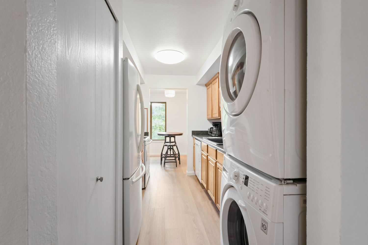 Narrow hallway with stacked washer and dryer leading to a bright kitchen with wood cabinets and light hardwood floors.