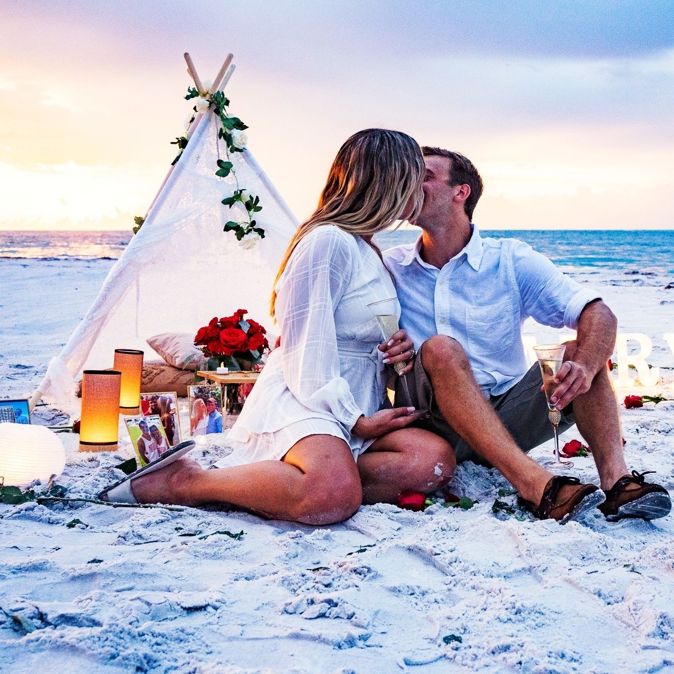 Romantic couple kissing at sunset beach picnic with white teepee, red roses, and candles on the sand.