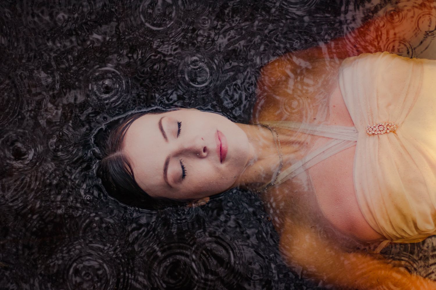 Woman with closed eyes floating in dark rippling water, wearing a light dress with auburn hair spread around her.