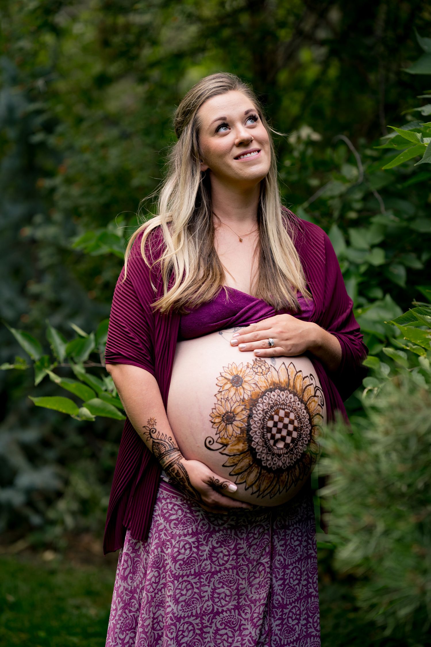 Smiling pregnant woman with henna belly art wearing burgundy wrap in a lush green garden.