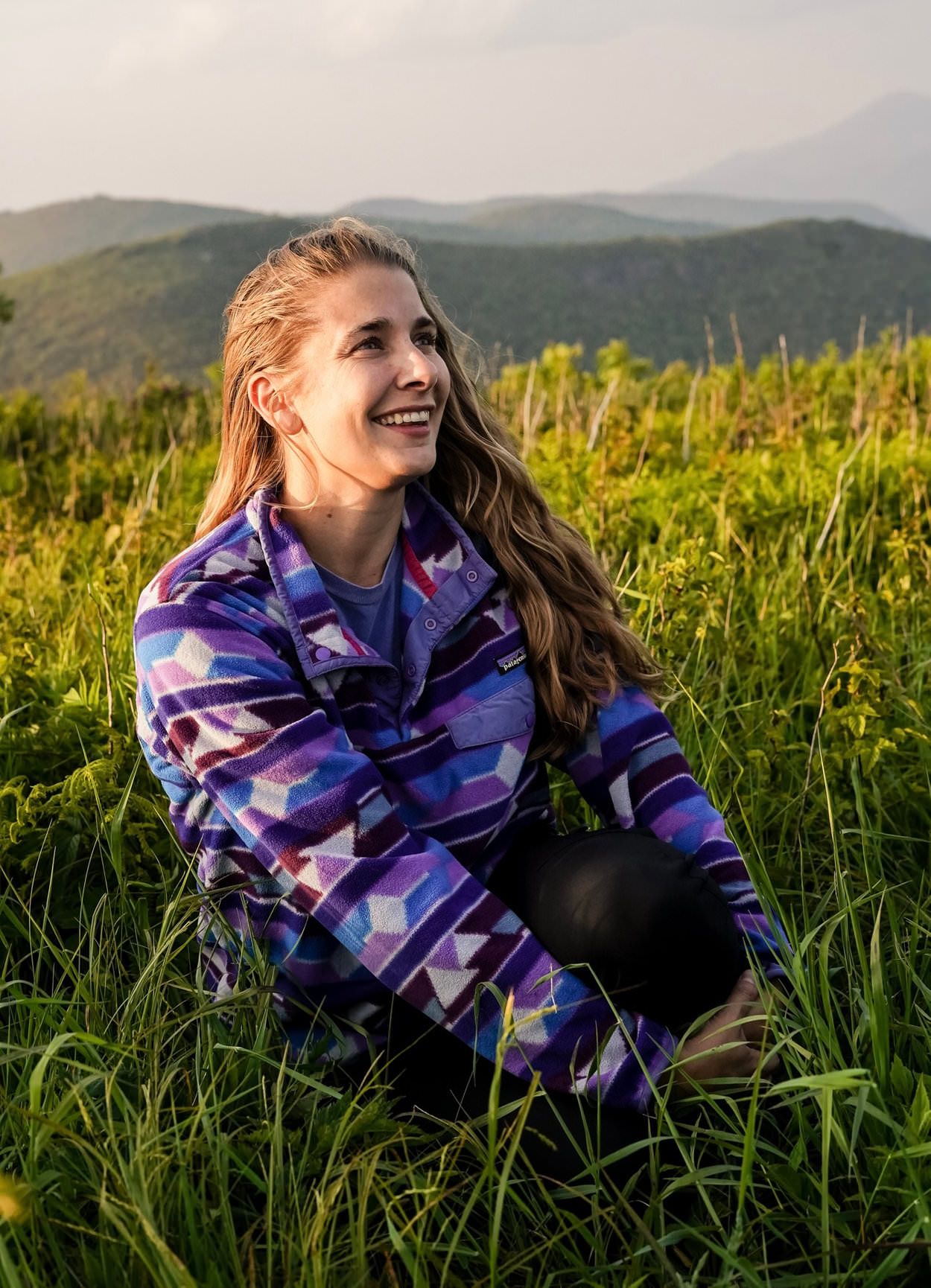 Smiling young woman in colorful Patagonia jacket sitting in green meadow with mountain backdrop.