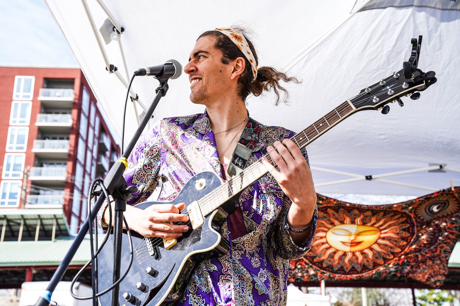 A man plays guitar in front of a bright sun tapestry during a live outdoor concert in downtown Boulder, filling the street with music.