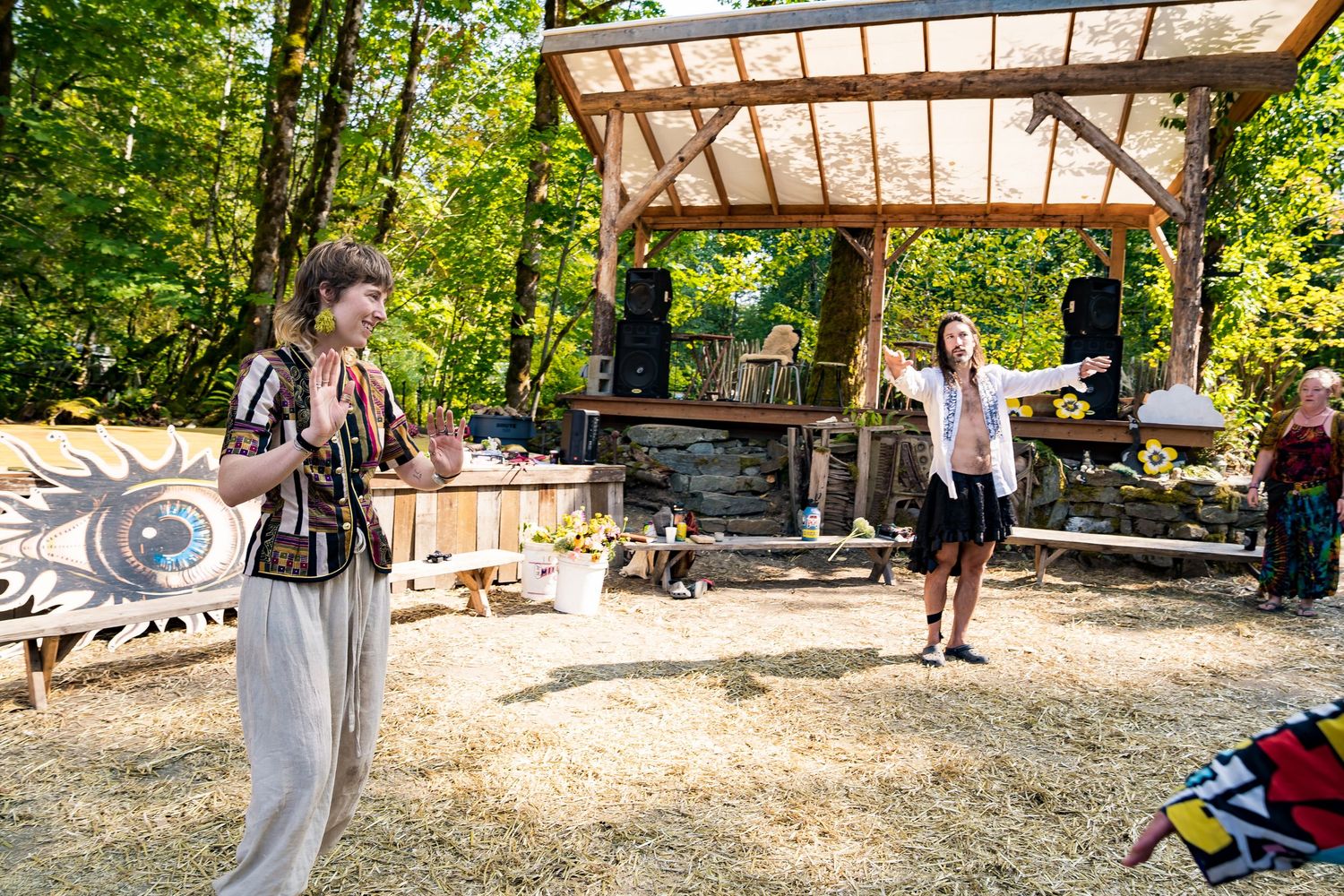 Two women stand in an outdoor woodland stage area surrounded by trees at a festival event.