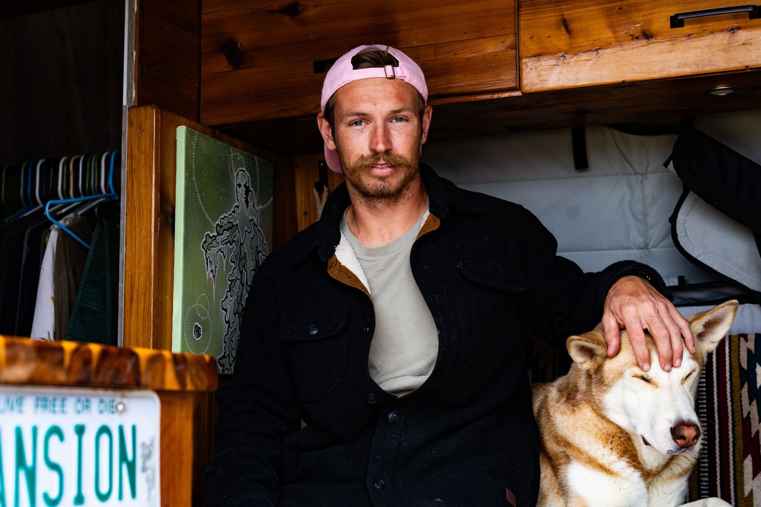 Man in black jacket and pink cap sits with a Shiba Inu dog inside a cozy van conversion.