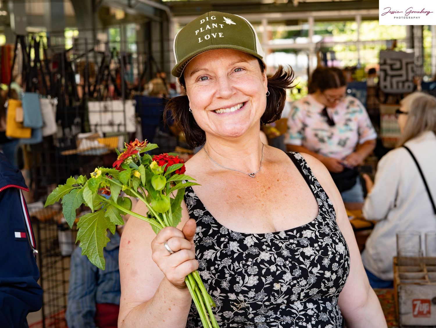 Smiling woman holding fresh red peppers with green stems at an outdoor farmers market.