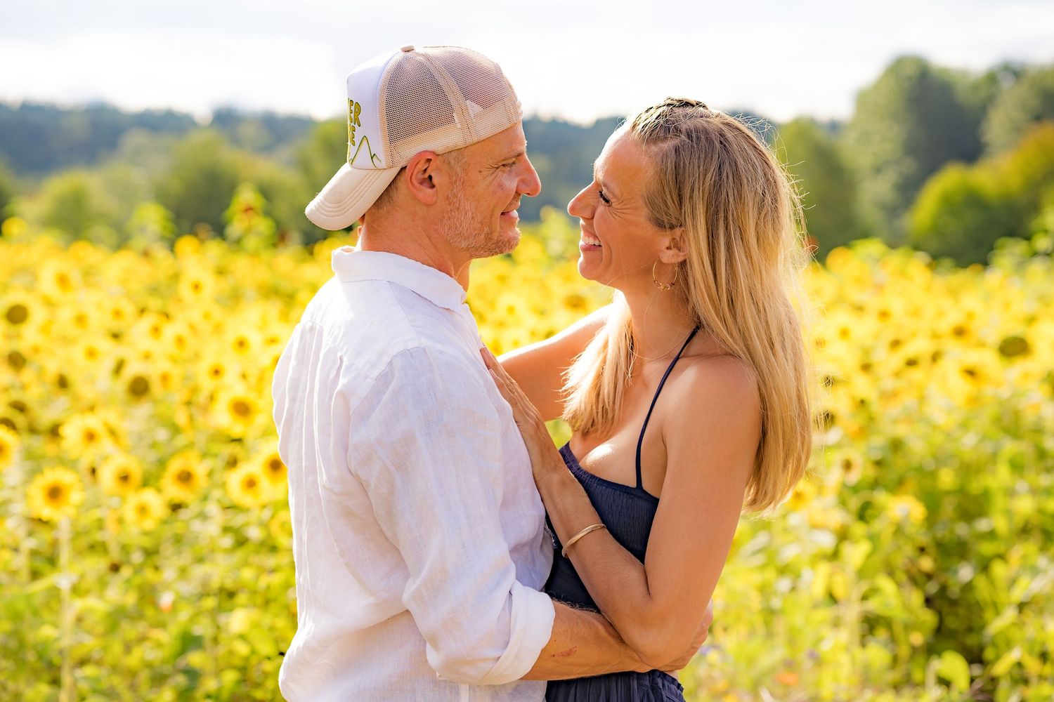 Couple embracing and smiling at each other in a vibrant sunflower field on a sunny day.