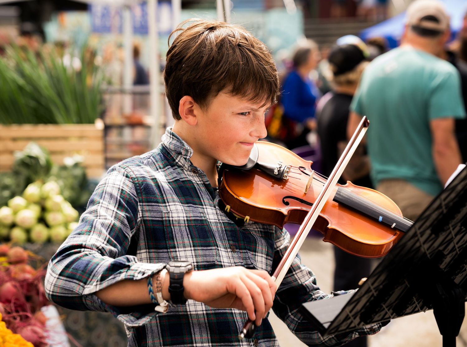 A young boy playing violin at an outdoor farmers market, wearing a plaid shirt, with produce visible in the background.