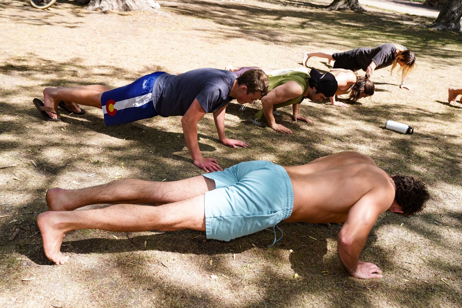 Group of people doing push-ups outdoors on dirt ground under shaded trees during a fitness workout session.