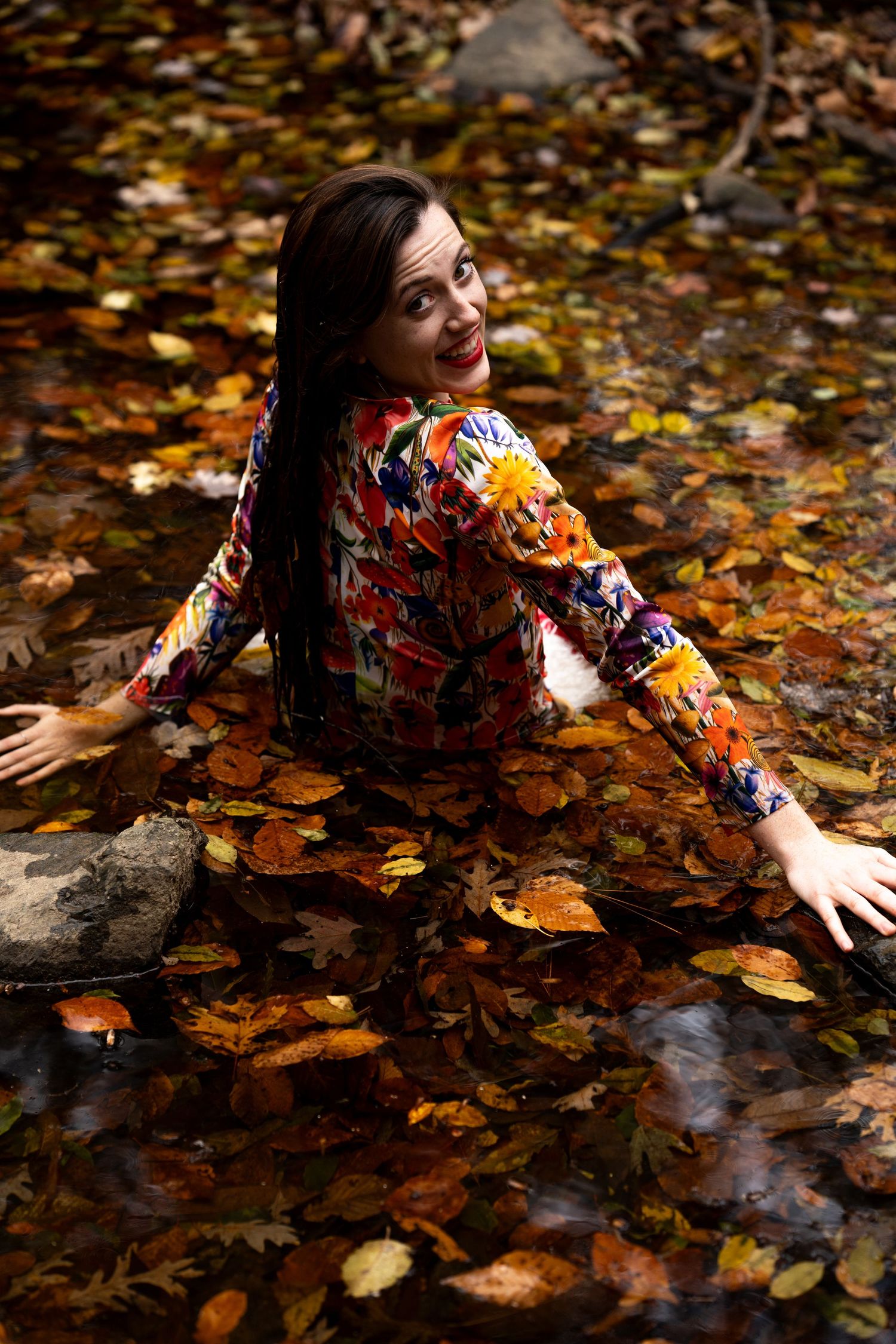 A person in a floral dress lies among scattered autumn leaves on the ground near water.