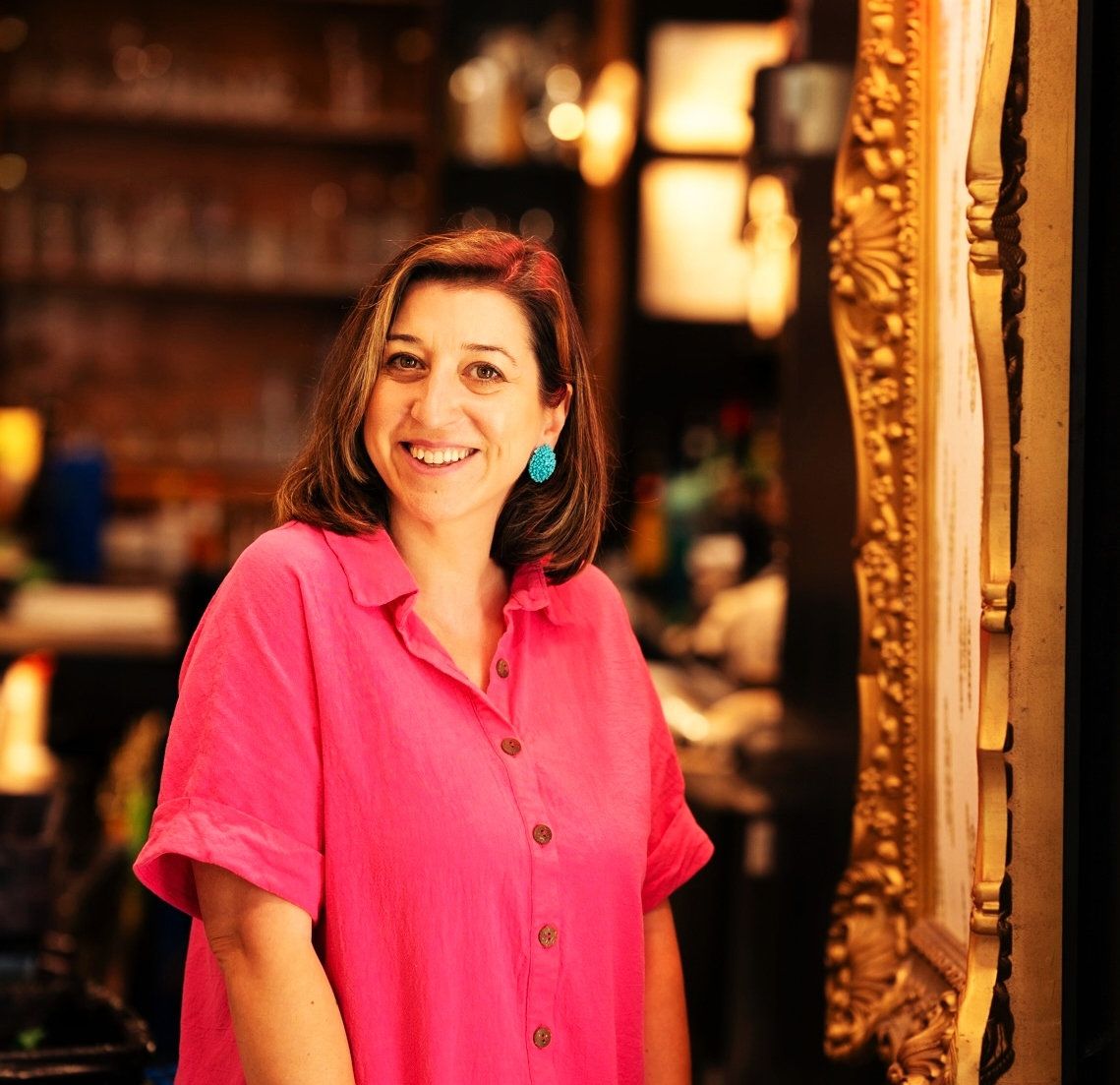 A photo of a woman wearing a bright pink button-up top, smiling into a mirror, wearing blue earrings.
