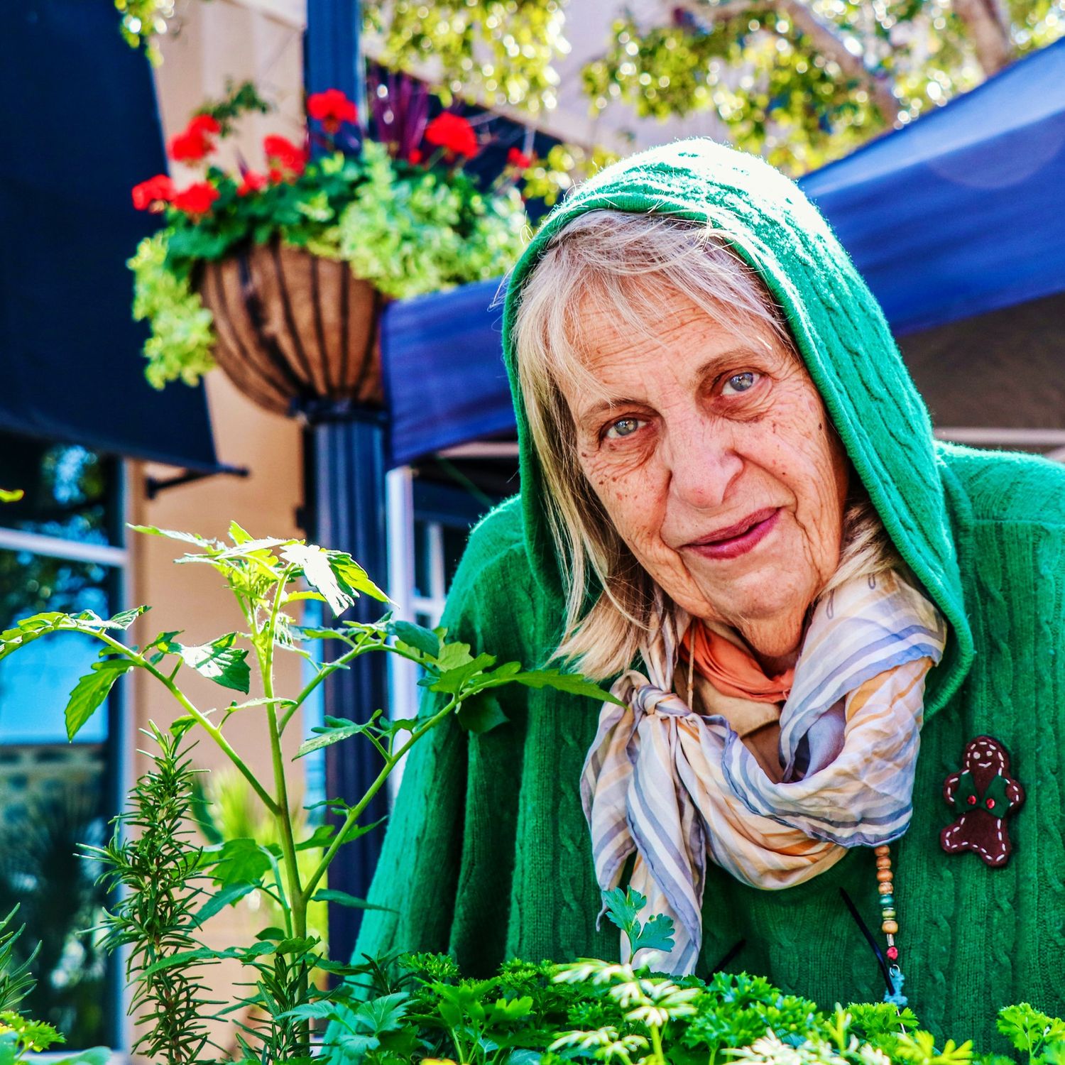 A photo of a woman wearing a green sweater, she is looking at the camera and there are red flowers behind her.