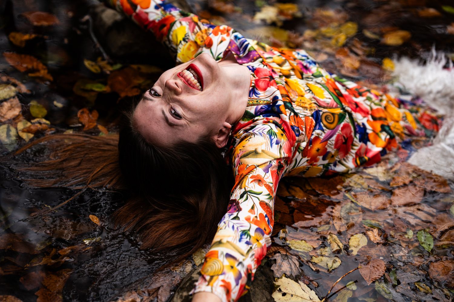 Woman in colorful floral dress lying among autumn leaves, laughing joyfully outdoors.
