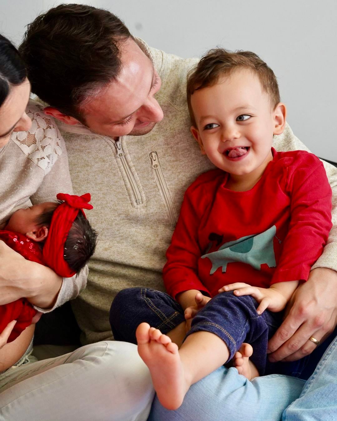 Family portrait with parents holding a newborn baby and a smiling toddler in a red Christmas sweater.