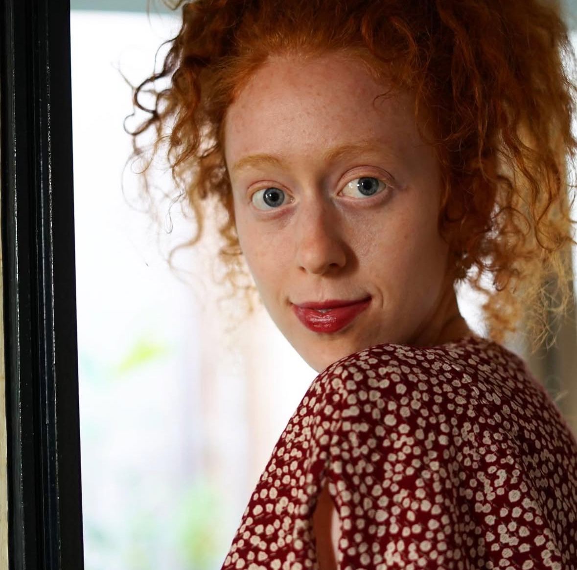 A young woman with curly red hair, blue eyes, and freckles wearing a red floral blouse stands near a window.