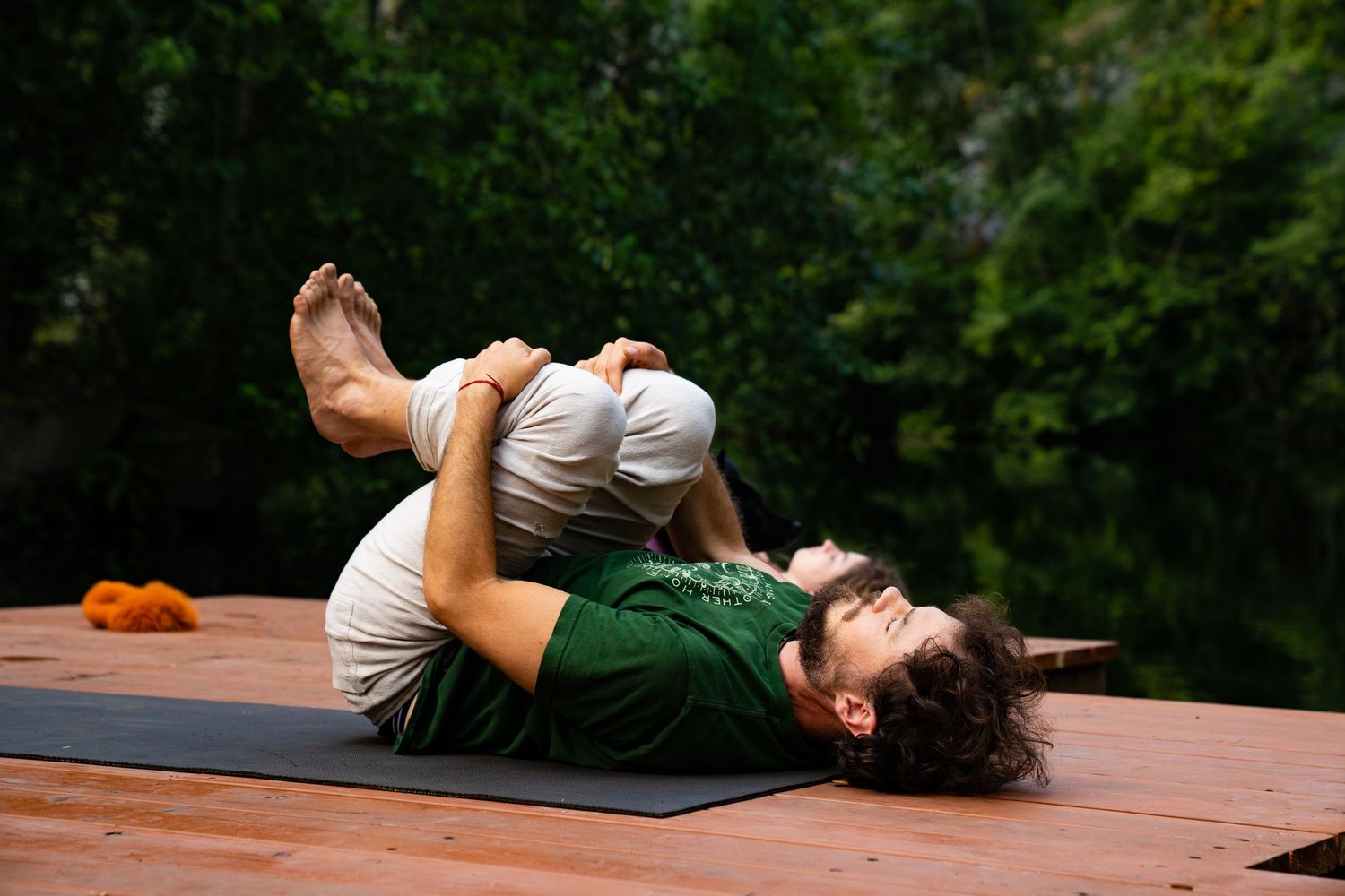 Man practicing advanced yoga pose on wooden deck outdoors, lying on back with legs folded overhead.