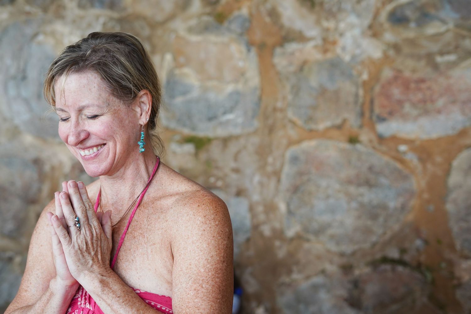Smiling woman in pink top practicing yoga namaste pose with hands pressed together near stone wall.