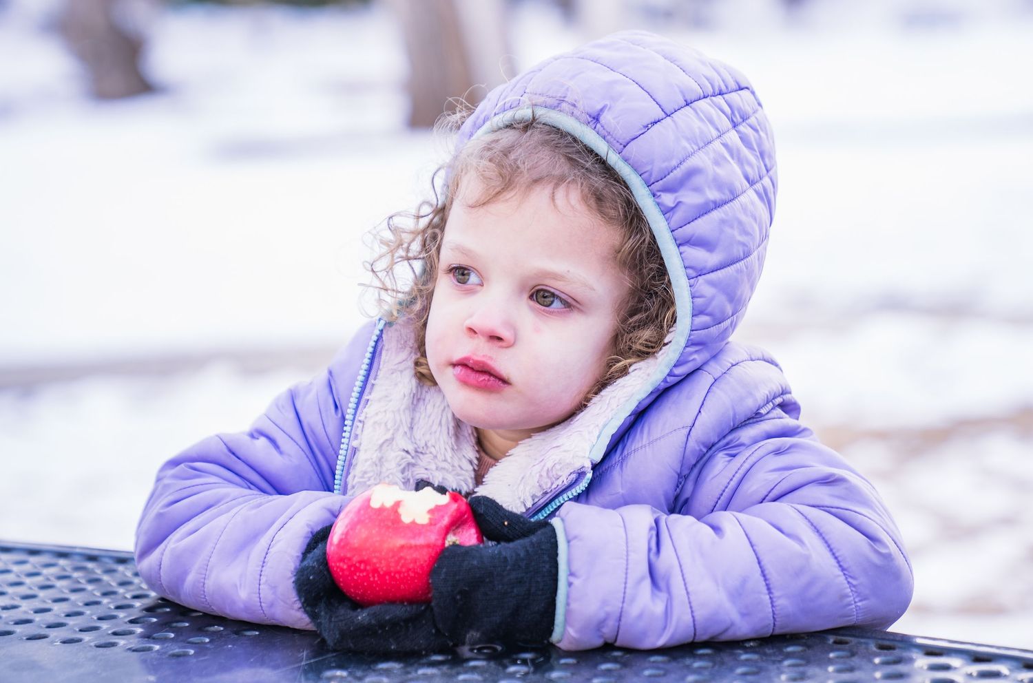 A child in a lavender winter coat lays on playground equipment while holding a red ball on a cold day.