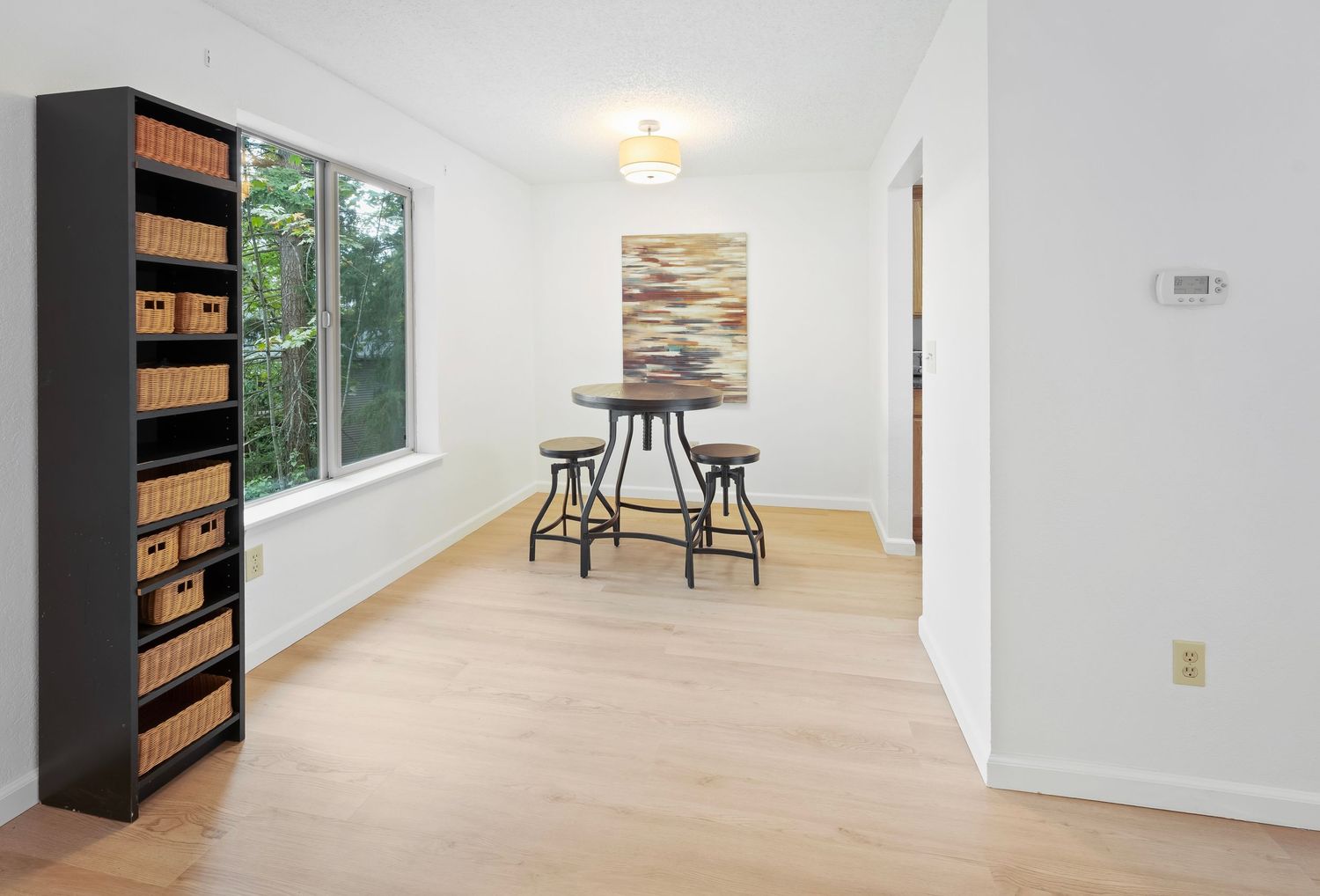 Bright dining nook with light hardwood floors, tall bookshelf with wicker baskets, round table with stools, and large window.