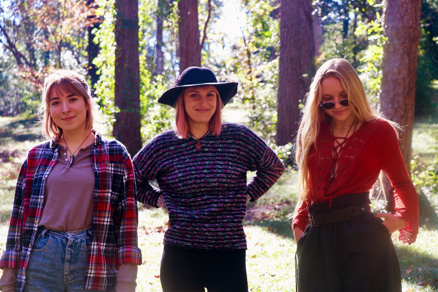 Three young women posing together in a sunlit forest, wearing casual flannel and knitwear outfits.