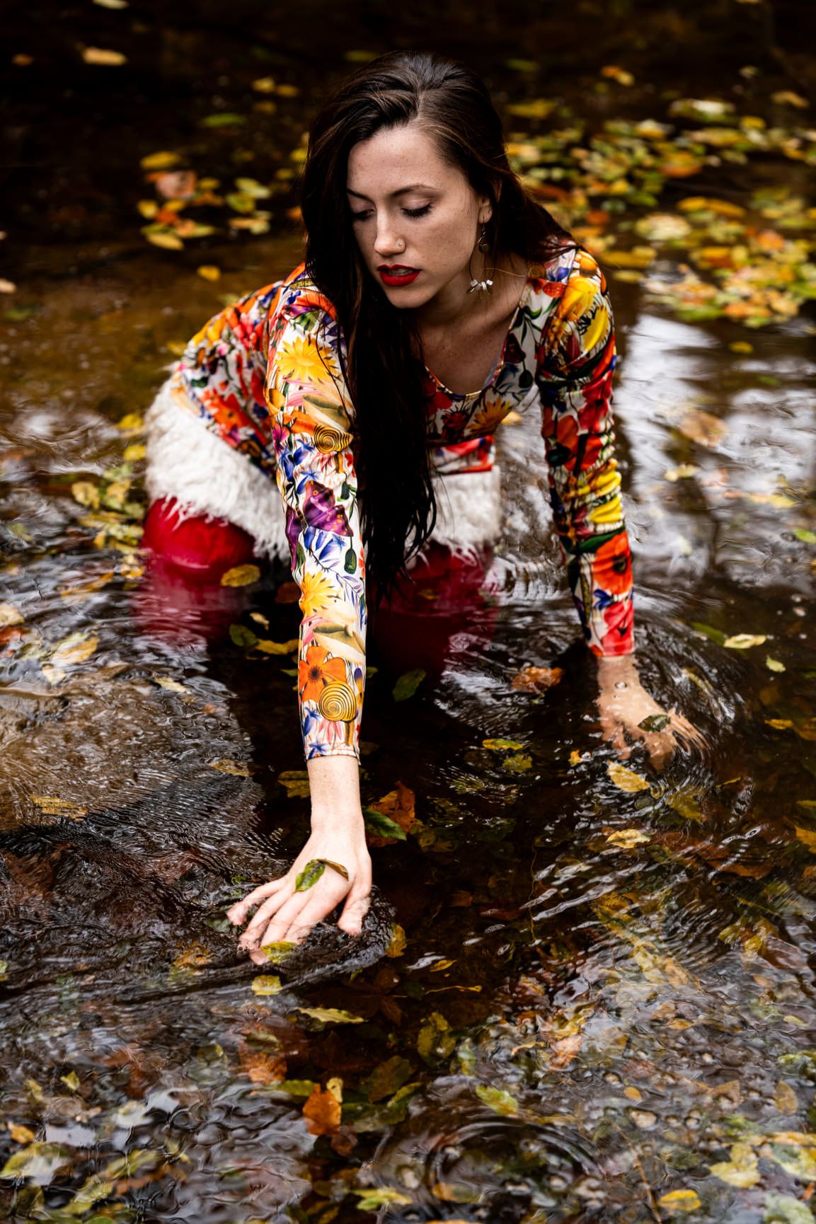 Woman in colorful floral outfit kneeling in shallow stream surrounded by autumn leaves.
