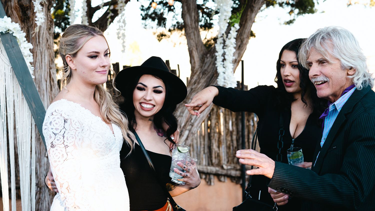 Four elegantly dressed people socializing outdoors at a festive event, including a bride in white and guests in stylish attire.