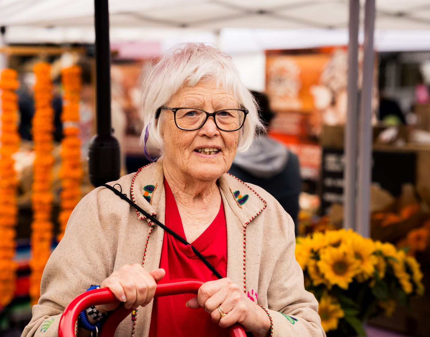 Elderly woman with white hair and glasses smiling at an outdoor farmers market with colorful flowers and produce.
