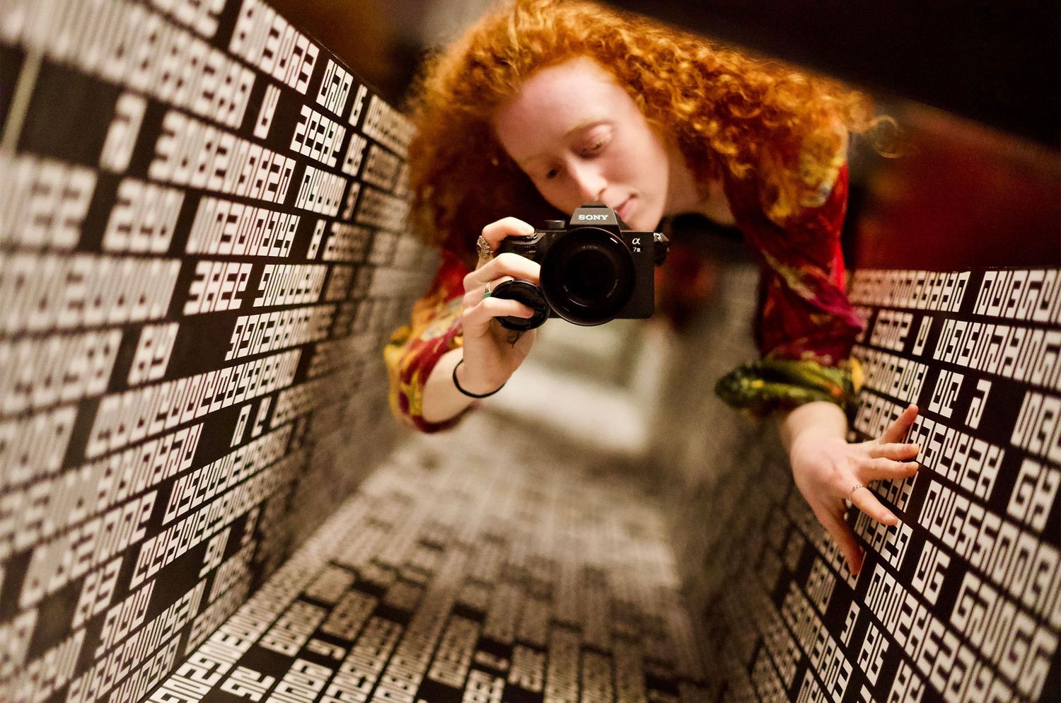Red-haired woman photographing herself in a mirror surrounded by walls covered in black and white text.