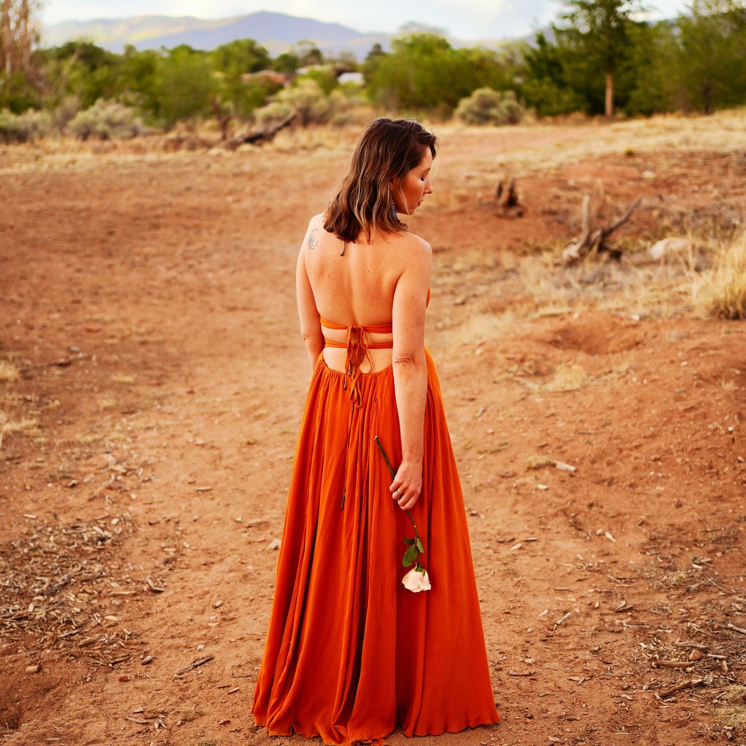 A photo of a woman wearing a flowy orange dress, she is standing on the dirt and is holding a rose behind her.