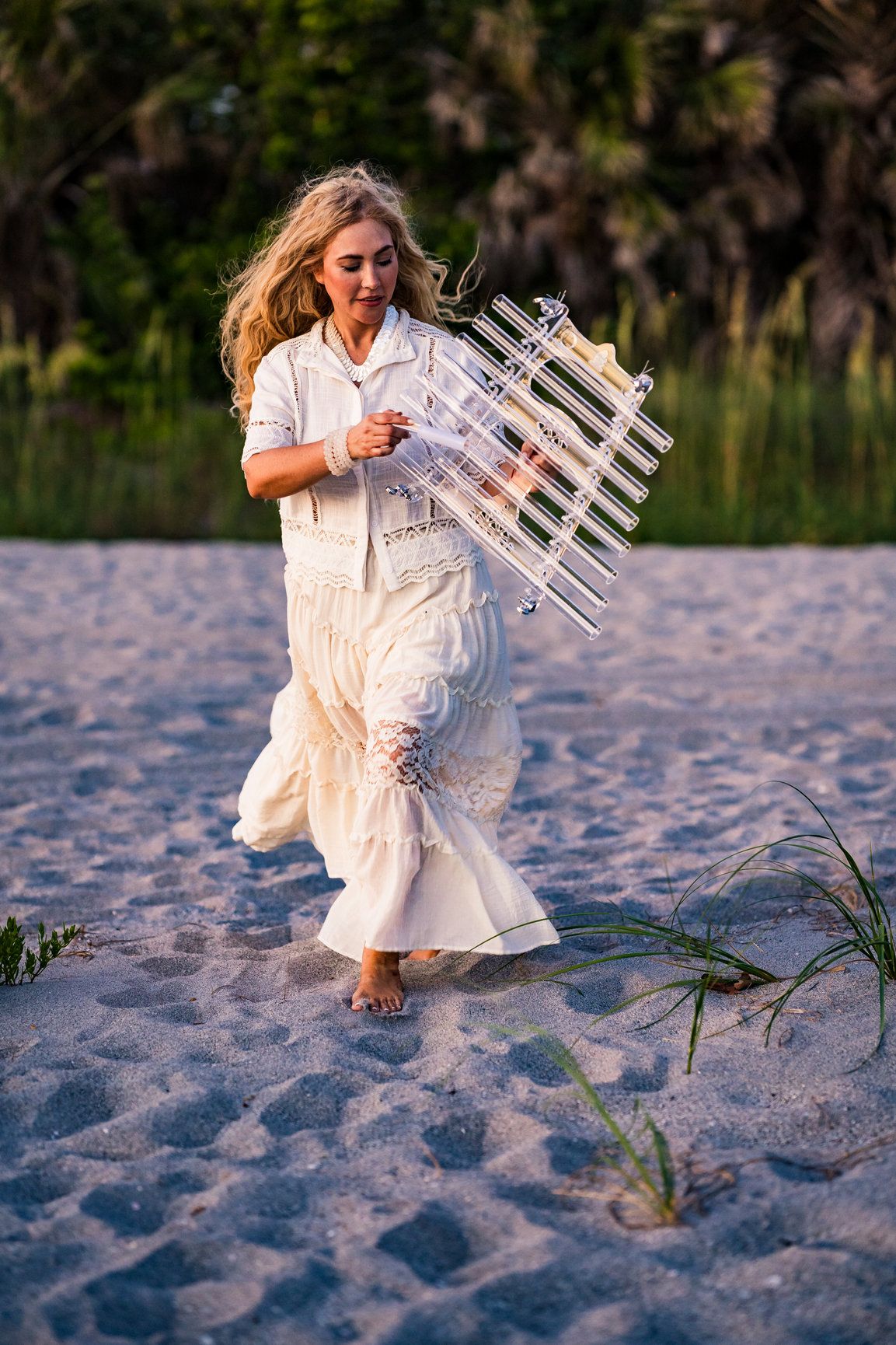 A photo of a woman walking on the beach, she has a white outfit and is playing an instrument while she is walking.