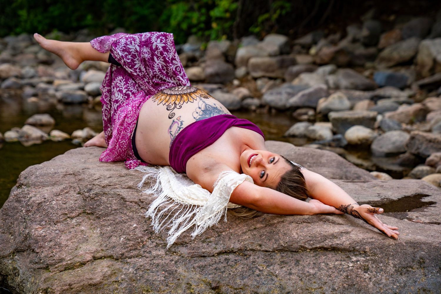 A person in a flowing purple dress lies gracefully across rocks near a stream creating an artistic pose.