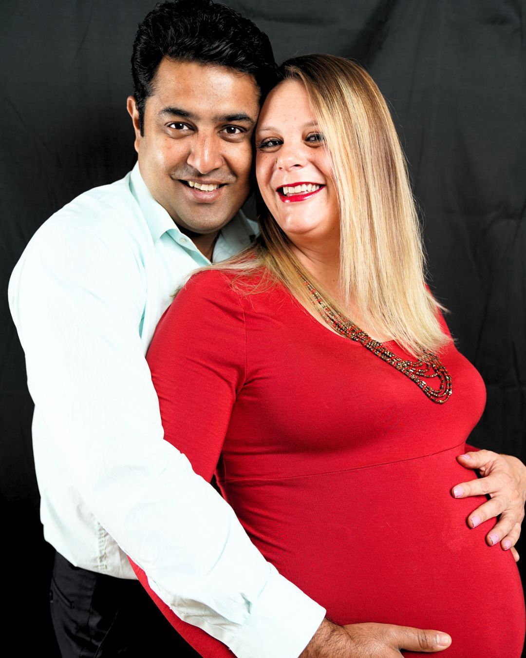 Smiling pregnant couple poses together, woman in red dress cradling baby bump against dark background.