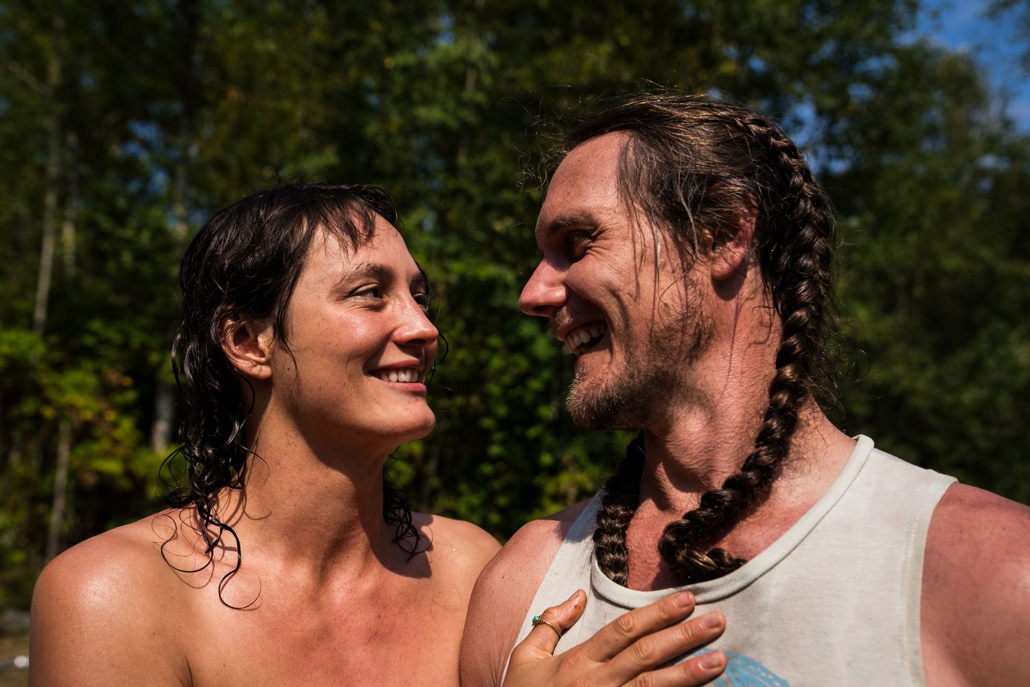 A smiling couple with wet hair gazes at each other outdoors among green trees on a sunny day.